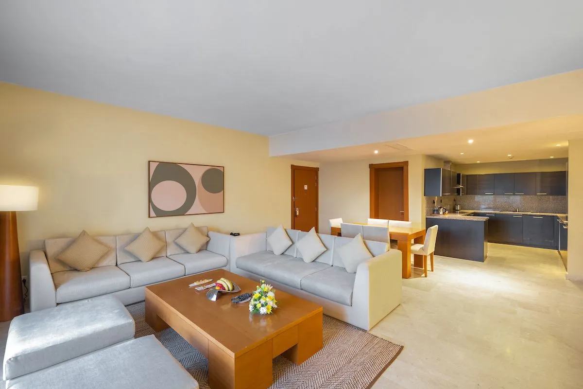 Bright and modern two-bedroom apartment living area with a gray sofa, wooden table, and kitchen in the background.