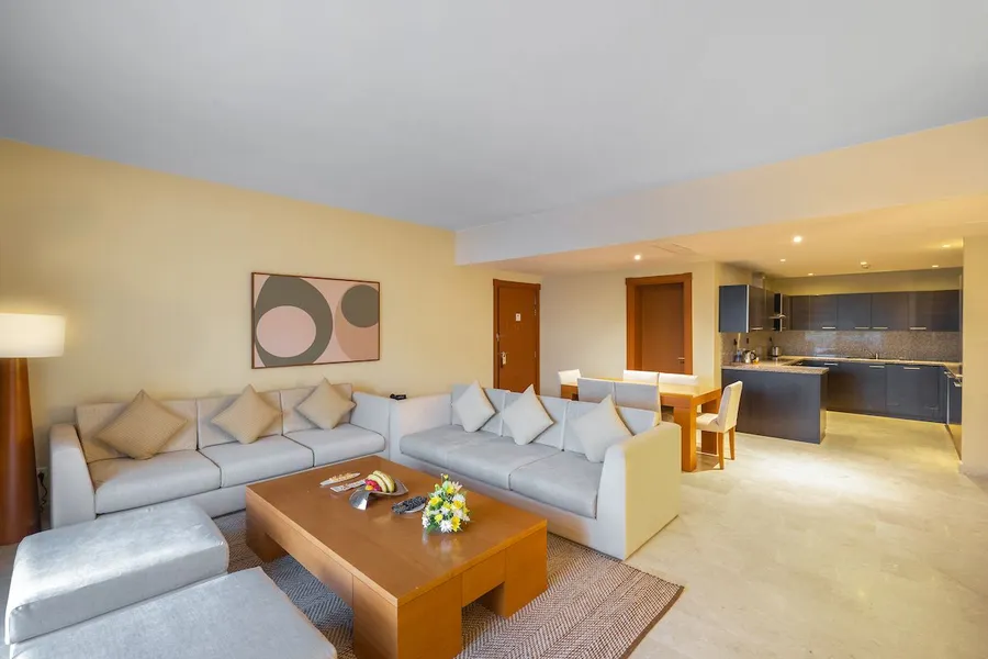 Bright and modern two-bedroom apartment living area with a gray sofa, wooden table, and kitchen in the background.